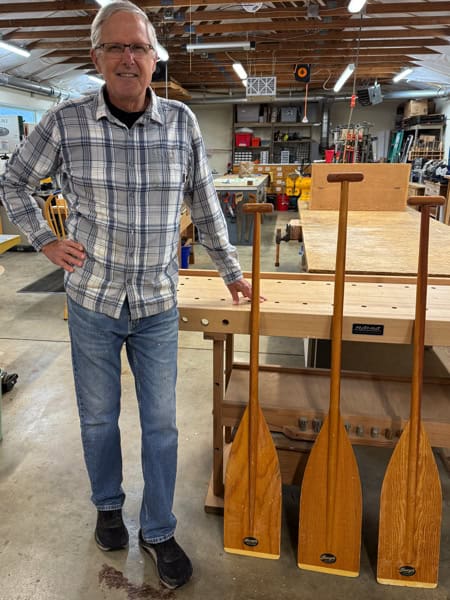 Man in plaid shirt standing next to three boat paddles